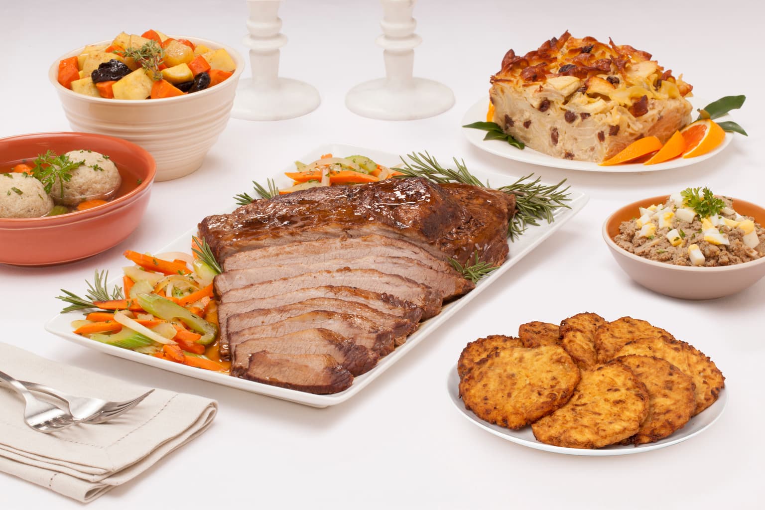 Partially sliced brisket with veggies on white platter surrounded by matzo balls in broth, tzimmes, noodle kugel, chopped liver and latkes. Napkin with forks next to the brisket.